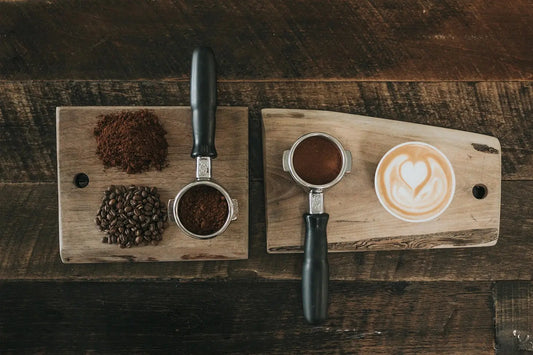 Café latte avec latte art en forme de cœur sur une planche en bois, accompagné de grains de café, de café moulu et de porte-filtres pour machine expresso.